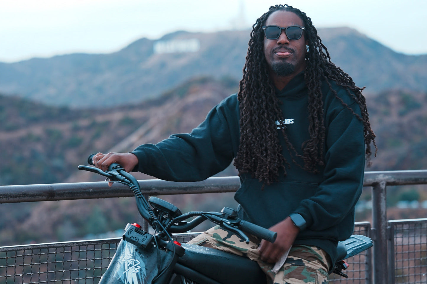 Man wearing sunglasses and hoodie sitting on a GT73 Pro electric motorbike with mountainous background
