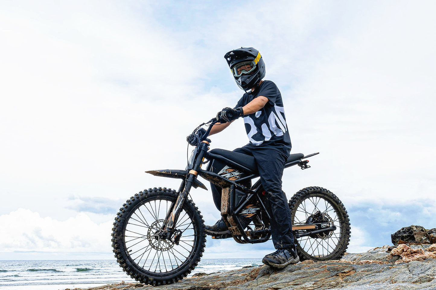 Rider on GT73 Pro electric motorbike with off-road tires on rocky terrain by the sea.