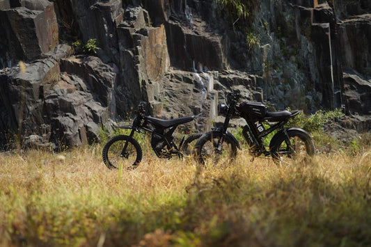 Two black electric bikes parked in a grassy area near rocky cliffs, ideal for beginner riders and off-road adventures