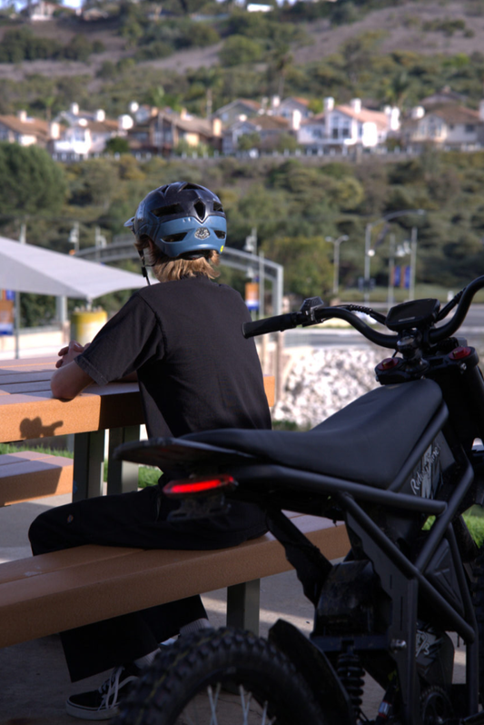 A rider in a helmet sits on the grass beside the Riding Times GT73 electric bike, with a scenic outdoor backdrop featuring trees and a residential area.