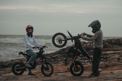 Two people with dirt bikes on a rocky beach with ocean waves in the background